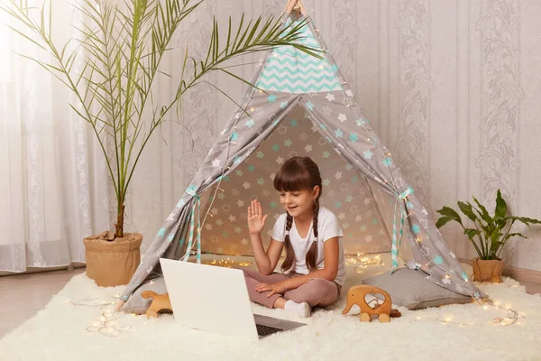 Portrait of cute little girl sitting in peetee tent and having video call or broadcasting livestream, looking at notebook screen, waving hand, saying bye or hello.