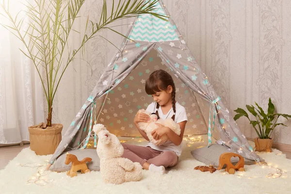 Indoor shot of little cute girl playing with soft toy teddy bear near the wigwam in the children's room, funny cute small female child having fun at home in peetee tent.