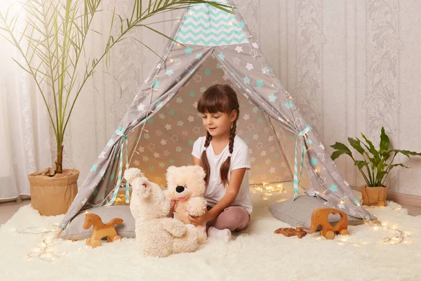 Indoor shot of cute girl playing with soft teddy bear and rabbit in wigwam, kid with pigtail wearing white t shirt sitting on floor on white carpet, having fun alone.