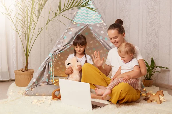 Indoor shot of happy family, mother elder daughter and toddler baby, sitting on floor near peetee tent and using laptop for talking on video call, waving hand, saying hello.