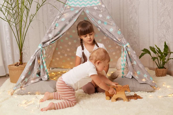 Indoor shot of little sisters playing in peetee tent with eco soft toy, preschooler girl and toddler baby sitting on floor near wigwam, having fun with interest.