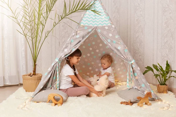 Indoor shot of little sisters playing in peetee tent with soft teddy bear, preschooler girl and toddler baby sitting on floor in wigwam, enjoying to spend time together.