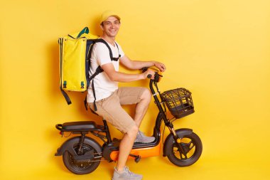 Kasket ve t-shirt giyen motosikletli bir teslimatçı, sarı arka planda izole edilmiş poz veriyor, kurye olarak çalışan bir adam, kameraya olumlu bir ifadeyle bakıyor..