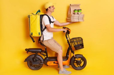 Kurye portresi, şapka ve tişört giyen elektrikli motosiklet süren, pizza dağıtan ve sarı arka planda izole edilmiş kahve getiren kurye portresi..