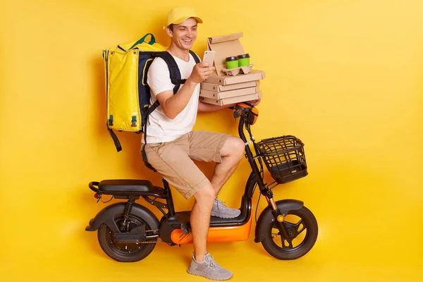 Young courier in white t shirt, pizza delivery man with thermo backpack and phone on moped isolated on yellow background. Fast transport express, home food delivery. Online order.