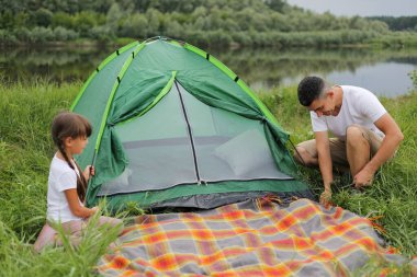 Portrait of young adult man wearing white t shirt setting up a tent in nature near lake or river, family having vacation together, picnic in open air, preparing for overnight stay in nature.