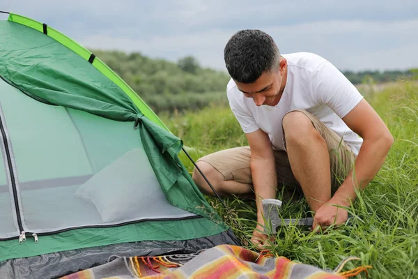 Full length outdoor portrait of hardworking man wearing white t shirt setting up a tent for overnight stay in nature near river, preparing everything for camping and picnic.