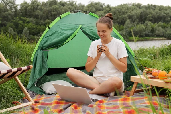 Attractive woman working on laptop near tent in nature, young freelancer sitting on the ground, relaxing in camping site near river. Remote work, outdoor activity in summer. Happy girl has a work on vacation.