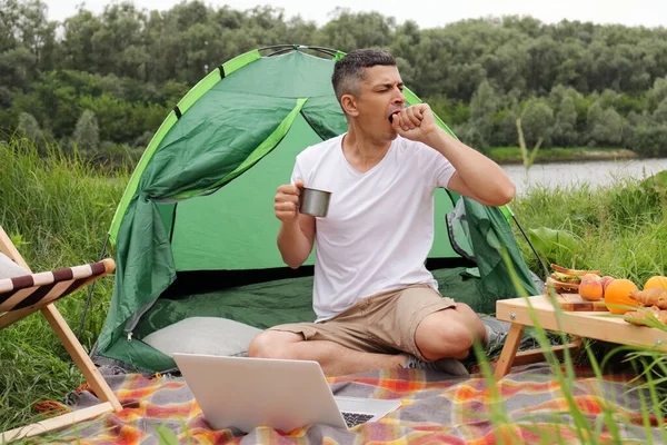 Tired male working on a laptop outdoors, freelancer has ability to work from anywhere in the world, sitting near tent and drinking coffee, needs energy, feels sleepy, yawning, covering mouth with hand