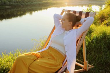 Horizontal shot of cute dark haired Caucasian woman wearing white shirt and yellow trousers sitting in the folding chair by the water, raised arms, looking at beautiful views.