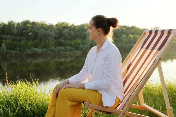 Side view portrait of young adult woman sitting in the folding chair at the picturesque bank of the river, enjoying the peacefulness of summer nature and watching the freedom of the deep.