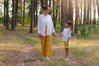 Outdoor shot of little girl standing with mom in the park, holding hand sand looking at each other, walking in beautiful summer forest, enjoying time together and nature.