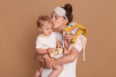 Indoor shot of loving mother standing with her infant daughter isolated over brown background, kissing her baby, expressing love and gentle, routine during maternity leave.