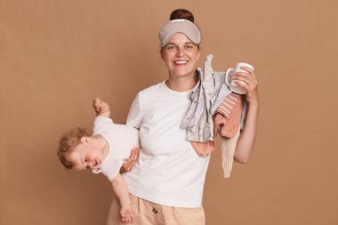 Horizontal shot of beautiful young woman wearing white t shirt and sleeping mask with baby in hands, standing looking at camera with charming smile, enjoying morning coffee.