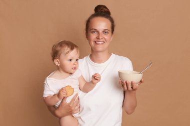 Horizontal shot of satisfied mother with bun hairstyle wearing white t shirt posing baby daughter in hands, looking at camera with toothy smile, holding plate with food for feeding child.