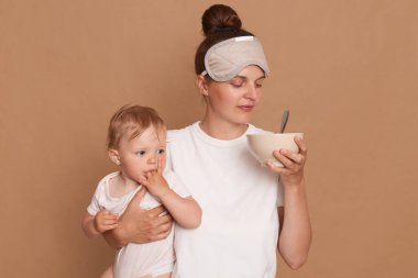 Portrait of woman wearing white t shirt and sleeping mask, holding infant kid in hands, smelling food in plate for feeding baby, posing isolated over brown background.