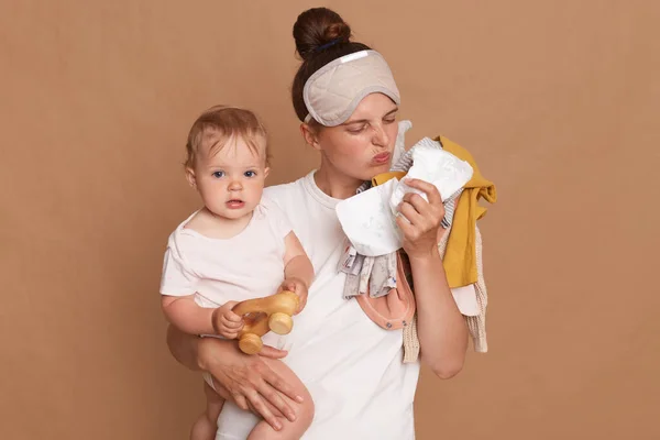 Young adult mother wearing white t shirt and sleeping mask, standing isolated over brown background, holding baby in hands and posing with lots of kid\'s clothing, smelling diaper, bad odor.