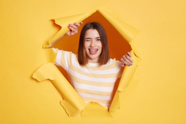 Indoor shot of funny childish woman wearing striped t shirt, winking and showing tongue out , expressing positive emotions, looking through breakthrough of yellow background.