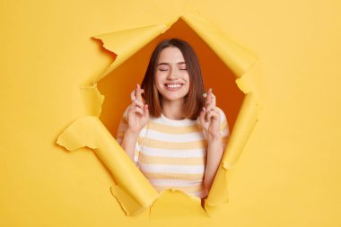 Image of hopeful optimistic woman wearing sttriped t shirt looking through breakthrough of yellow background, standing with crossed fingers, making wish.