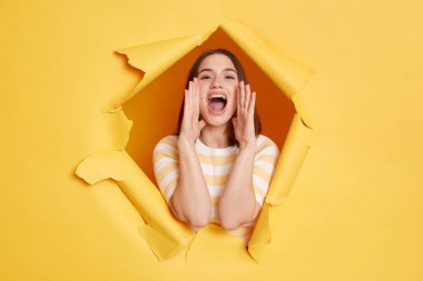 Horizontal shot of positive woman wearing striped t shirt posing standing through yellow paper torn hole, screaming, announcing advertisement with optimistic expression.