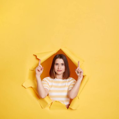 Indoor shot of dark haired young adult woman wearing striped t shirt looking through yellow paper torn, pointing fingers up, looks at camera, copy space for advertisement.
