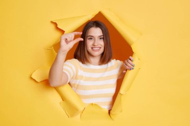 Woman wearing striped t shirt looking through breakthrough of yellow background, showing a little bit gesture, inch or centimeter, disappointed with minimum size.