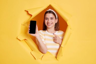 Portrait of smiling positive woman wearing striped t shirt showing cell phone with empty display for advertisement or promotion, posing standing through yellow paper torn hole.