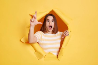 Horizontal shot of astonished surprised woman wearing striped t shirt posing standing through yellow paper torn hole, looking at camera with widely opened mouth.