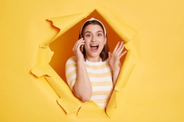 Indoor shot of excited amazed woman wearing striped t shirt looking through breakthrough of yellow background, talking phone with her friend, gossiping.