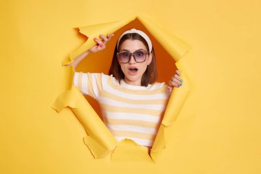 Portrait of amazed woman wearing striped t shirt looking at camera with widely opened mouth, expressing astonishment, sees something shocked, standing through yellow paper torn hole.