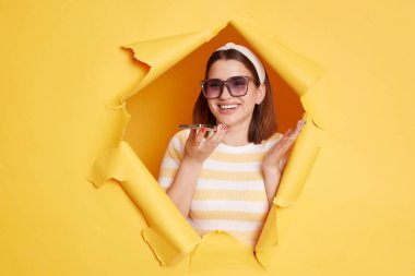 Image of smiling delighted woman wearing striped t shirt, recording voice message for friend or using mobile assistant, looking through breakthrough of yellow background.