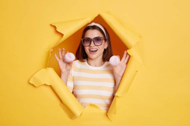 Portrait of attractive young dark haired woman wearing hair band and striped t shirt standing in yellow paper hole, holding marshmallows in hands, being amazed.