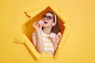 Image of woman Caucasian wearing hair band and striped t shirt, biting marshmallow, being hungry, being happy, eating delicious sugary dessert, standing in yellow paper hole.