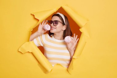 Horizontal shot of satisfied delighted woman wearing hair band and striped t shirt standing in yellow paper hole, smelling marshmallow and wants to eat sugary dessert.