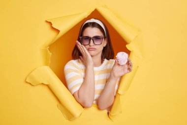 Portrait of unhealthy sick woman wearing hair band and striped t shirt standing in yellow paper hole, feeling pain after eating sugary dessert, touching her cheek.