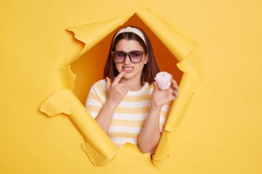 Portrait of Caucasian woman wearing hair band and striped t shirt felling discomfort in her teeth, pointing, holding marshmallow, standing in yellow paper hole.