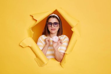 horizontal shot of hungry positive woman wearing hair band and striped t shirt holding sugary marshmallow and showing tongue out, standing in yellow paper hole.