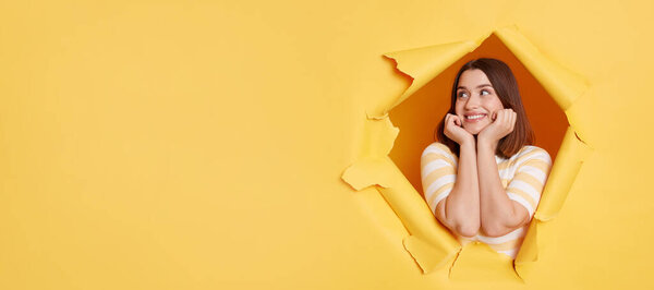 Smiling delighted woman wearing striped t shirt, expreressing positve emotions, copy space for advertisement or promotional text, posing standing through yellow paper torn hole.