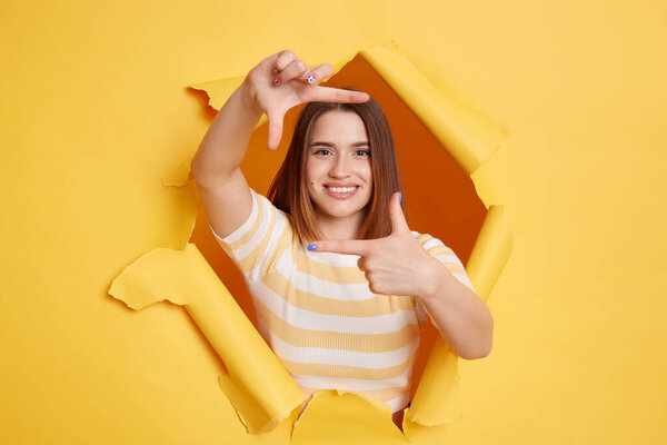Внутренний снимок улыбающейся женщины в полосатой футболке, смотрящей сквозь прорыв жёлтого фона, жестикулирующей фотографии, фокусирующейся на камеру, наблюдающей со счастливым выражением лица