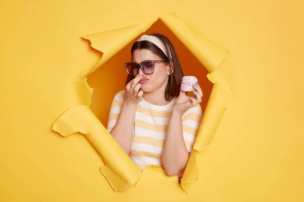 Indoor shot of young dark haired woman wearing hair band and striped t shirt standing in yellow paper hole, holding marshmallow and pinching her nose, bad odor.
