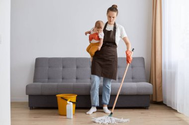 Image of sad upset tired woman wearing brown apron with mop and little toddler daughter in hands cleaning in living room, looking down with sadness, needs rest.