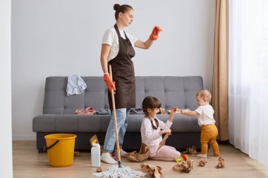 Indoor shot of exhausted sad upset woman with bun hairstyle wearing brown apron looking at mess on the floor after cleaning room, looks at her children sitting on floor and throwing paper.
