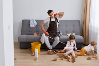 Horizontal shot of tired exhausted man with terrible pain in neck, sitting on sofa and touching back, having rest while washing floor in room where his children playing.