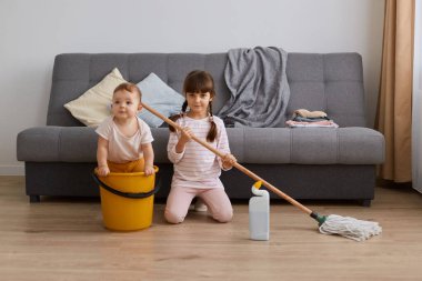 Indoor shot of little girl with mop in hands and her infant sister doing household the living room together, having fun while tidying up the apartment, baby sitting in bucket.
