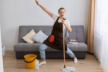Image of happy young woman singing while washing floor in living room, using wet moist mop as microphone, having fun during house chores, enjoying domestic duties, gamifying cleaning routine