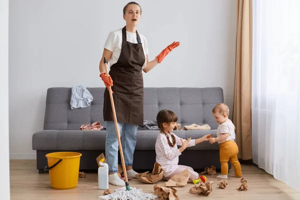 Portrait of tired mother wearing brown apron cleaning at home, woman with mop cleaning with kids sitting on floor at the sofa, exhausted housewife of tidying up house.