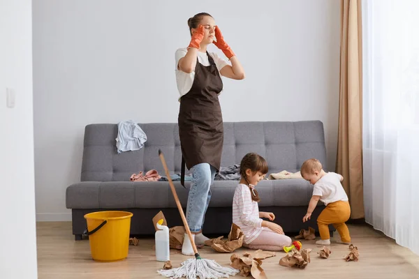 Image of tired mother standing near her daughters, mommy cleaning at home, woman touching head, kids sitting on floor and doing mess, exhausted housewife.
