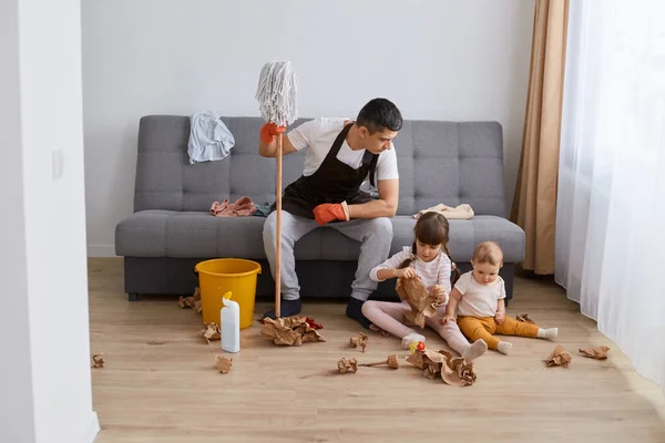 Indoor shot of man washing the floor with a mop at home, sitting on sofa to rest and talk with his daughter who playing on the floor with paper, father looking at kids with serious expression.