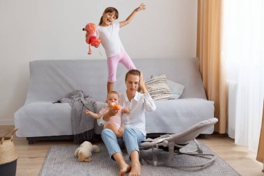 Indoor shot of exhausted dark haired young adult woman sitting on floor near sofa and keeping hand on her head, being tired of noisy little kids, wearing white t shirt and jeans.