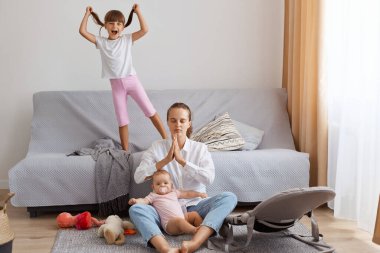 Indoor shot of dark haired Caucasian young adult woman wearing white shirt and jeans trying to calm down while spending time with her little children, keeps palms together in praying gesture.